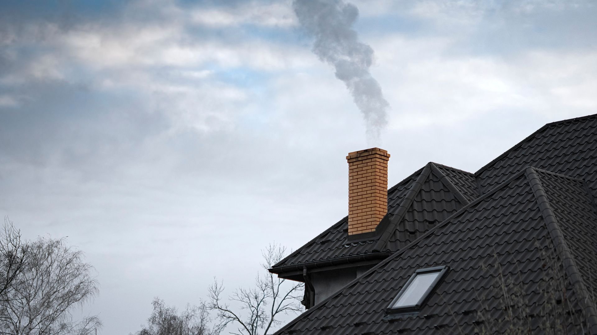 Smoke rising from brick chimney on dark tiled roof against cloudy sky