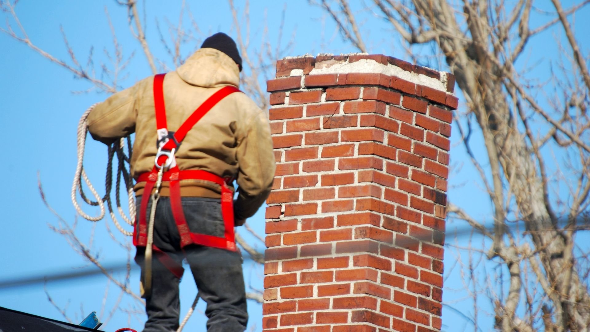 Chimney sweep in safety harness working on brick chimney against blue sky